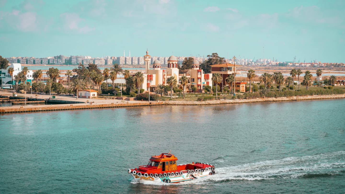 Makadi Bay, Egypt - Scenic view of a red boat cruising near a city with palm trees and vibrant architecture.