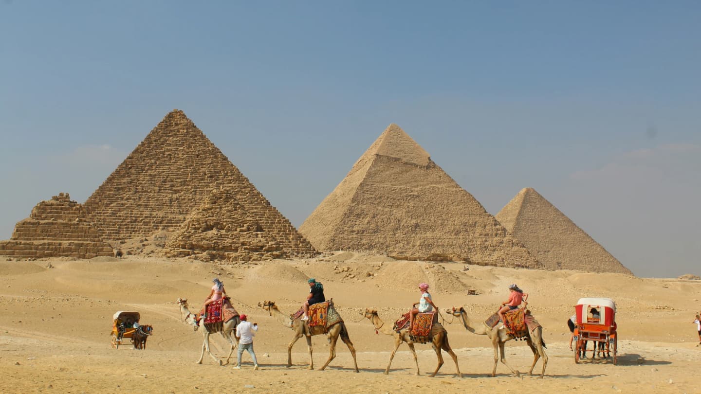 Safaga, Egypt - People riding camels past pyramids under a clear sky.