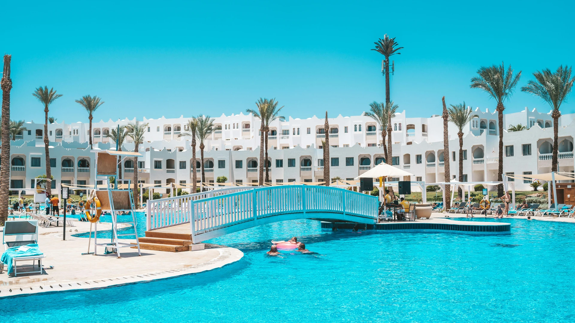 El Alamein, Egypt - Scenic view of a resort pool with a footbridge and palm trees under a clear sky.